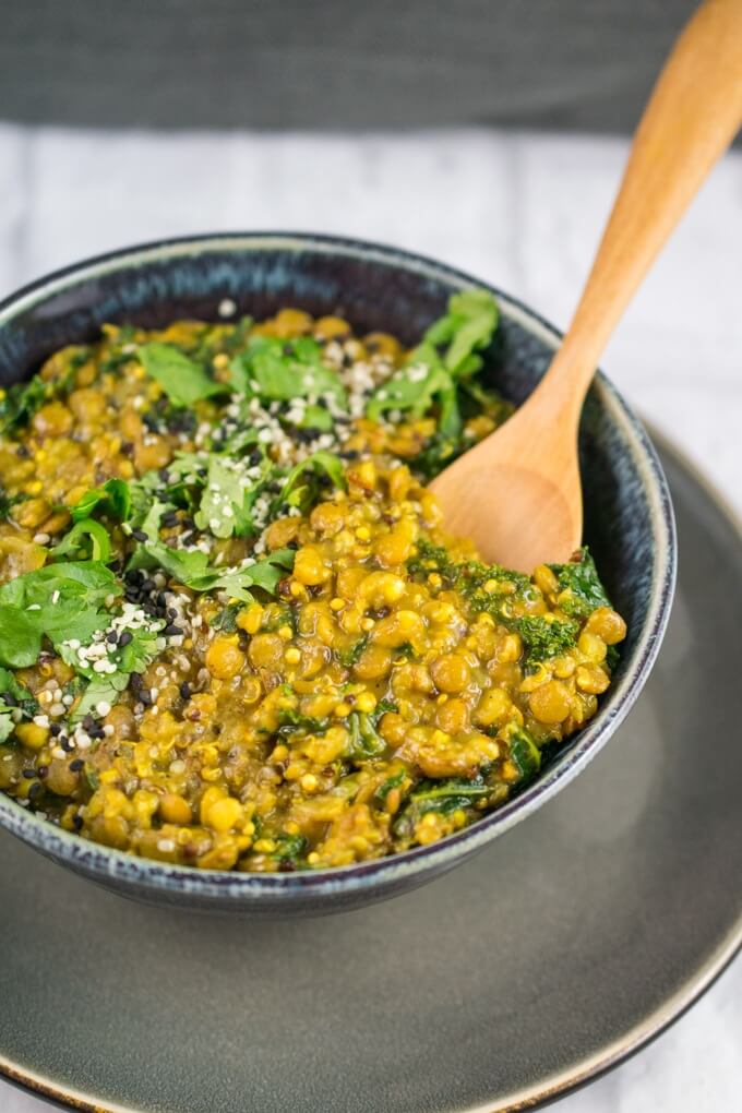 A spoonful of creamy one pot curried lentils and quinoa being scooped out of a blue ceramic bowl with a gray plate and white wooden table.
