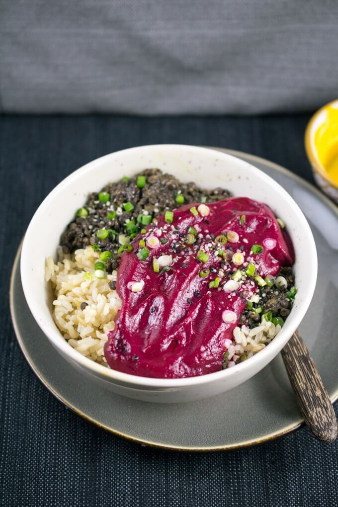 Overhead view of a bowl of Peruvian lentils, burgundy beet sauce, brown rice, garnished with scallions, hemp seeds, and black sesame, on a gray plate with a small bowl of chili sauce in the background.
