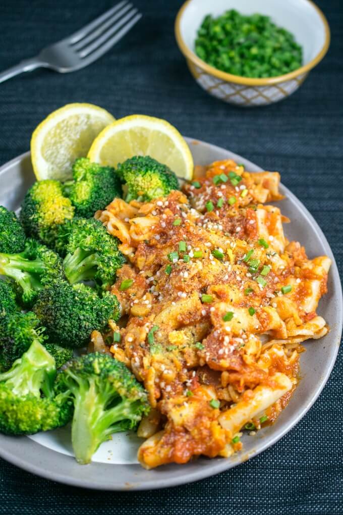 A plateful of penne pasta with a bright red chickpea and roasted red pepper sauce, steamed broccoli and lemon wedges