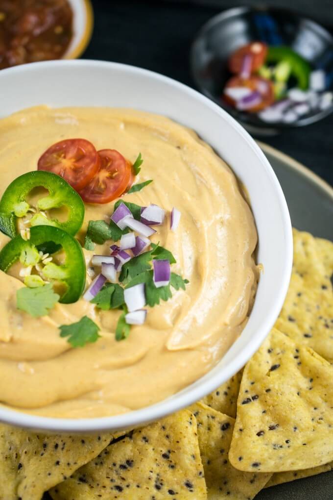 Close-up of dairy-free nacho cheese dip, made with silky pureed cauliflower. With vegetable toppings, tortilla chips, and jalapeno peppers!