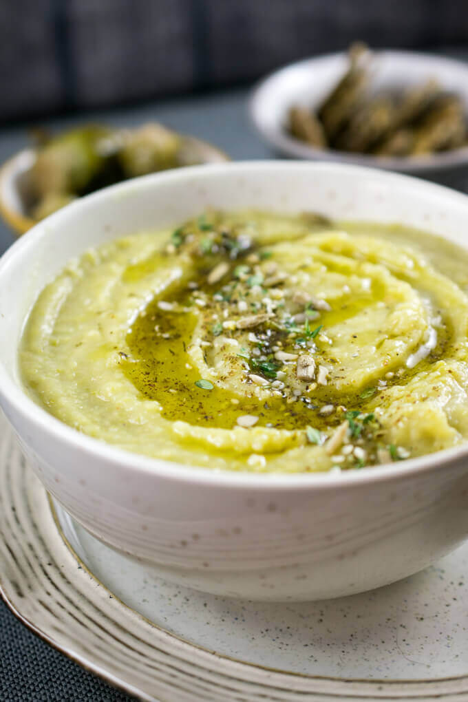 A close-up shot of creamy and light Middle Eastern green split pea soup, with a smooth but thick pureed texture, with bowls of Brussels sprouts and seeded crackers in the background