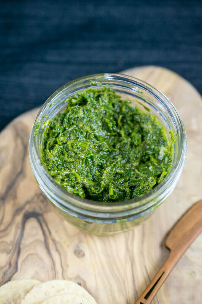 View into a glass jar with green carrot top pesto atop a wooden cheese board next to a cheese knife