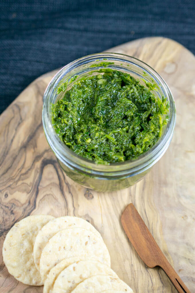 A jar of bright green carrot top pesto on a cheese platter, with flecks of carrot greens, mint, and walnuts visible.