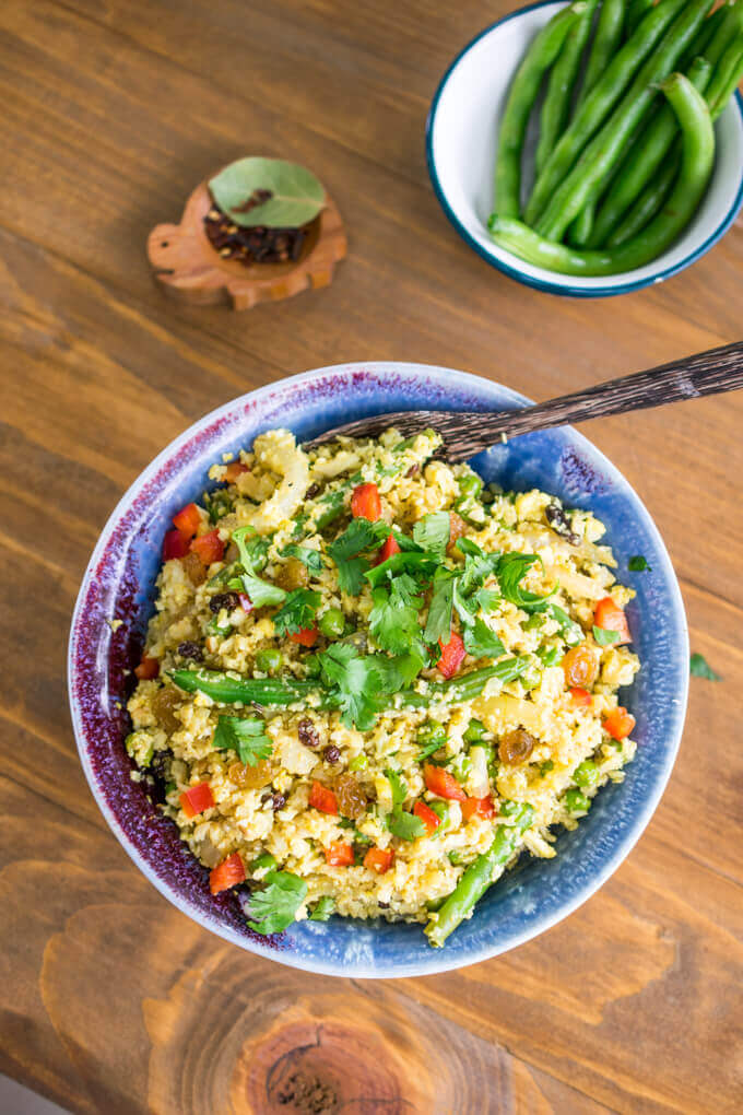 Overhead view of Indian curried cauliflower rice biryani next to a bowl of fresh green beans and a bowl of red chili flakes