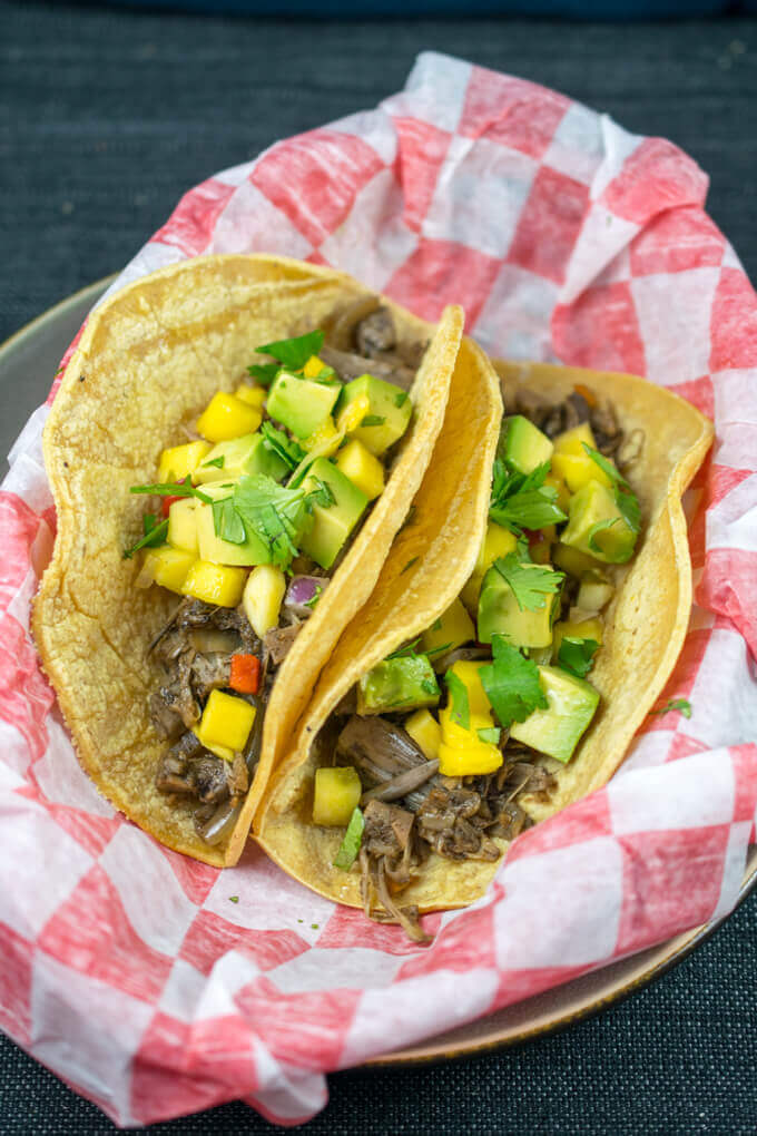 Two Jamaican jerk jackfruit tacos in a lined basket, with folded corn tortillas, jerk pulled jackfruit, and topped with avocado and mango