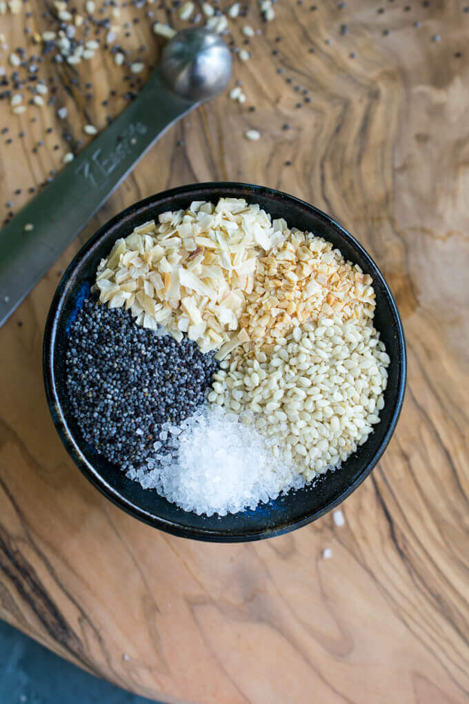 Overhead view of the components of everything bagel seasoning in a ceramic dish: minced onion, minced garlic, poppy seeds, coarse salt, and sesame seeds.