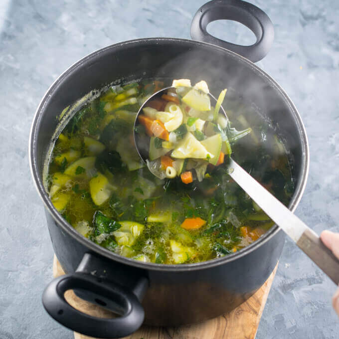 A pot of vegan summer minestrone soup with a ladle being scooped out that shows macaroni pasta, carrots, spinach, and zucchini