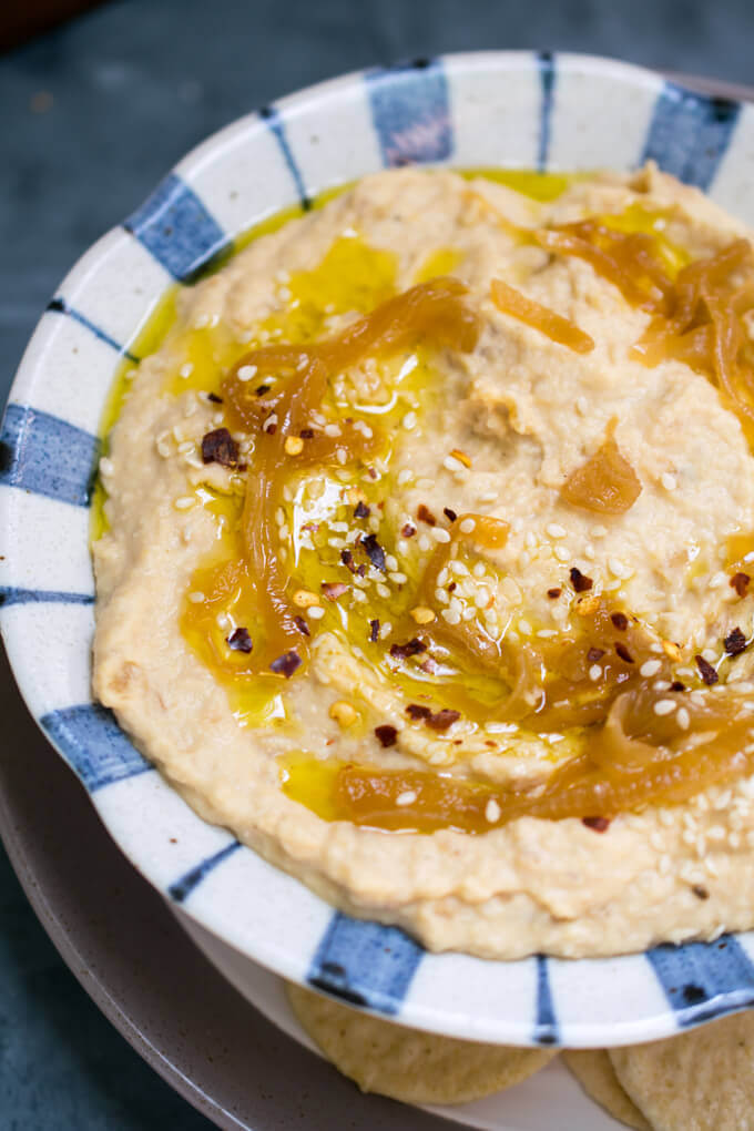 A bowl of caramelized onion hummus garnished with sesame seeds, red pepper flakes, and extra virgin olive oil