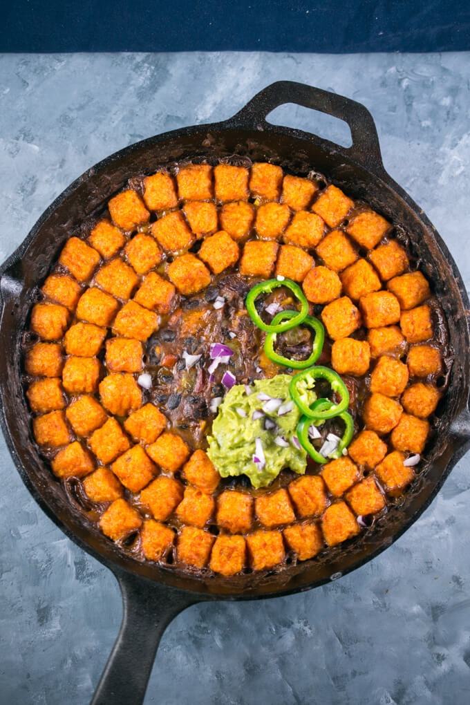 Overhead view of a sweet potato hotdish with a creamy black bean layer layered with sweet potato tater tots, with a dollop of guacamole and 4 sliced jalapenos on top