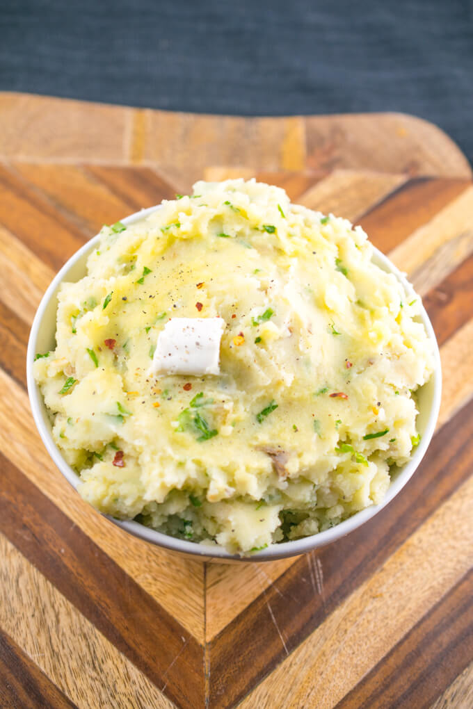 A cutting board with a bowl of rich vegan mashed potatoes with flecks of green parsley and red pepper flakes mixed in.