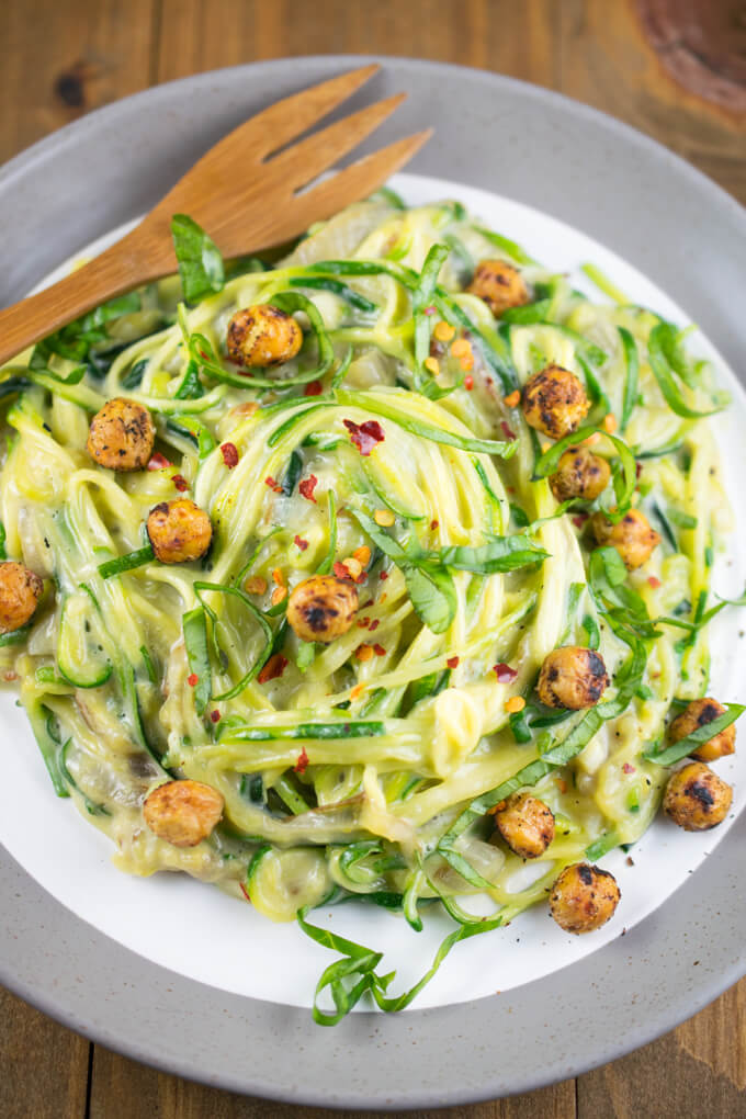 Overhead view of a pile of creamy garlic zucchini noodles on a plate, garnished with spice crusted chickpeas and red pepper flakes, next to a wooden fork.