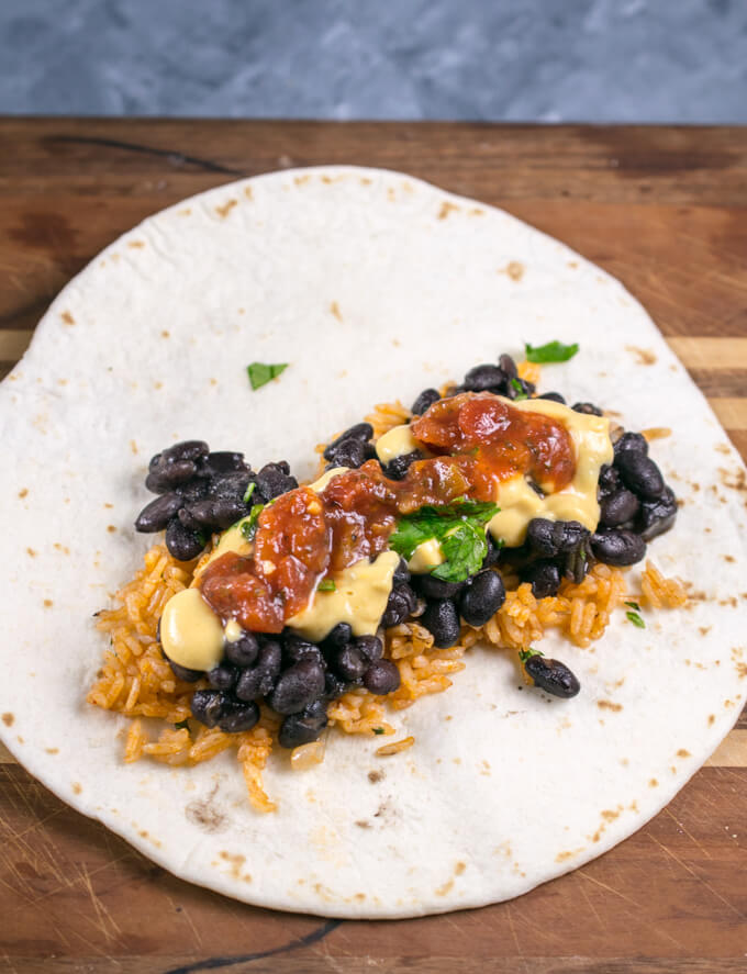 Vegan burrito filling on a tortilla before the burrito has been rolled up. Showing layers of Mexican rice, black beans, dairy-free queso, and a little bit of tomato salsa.