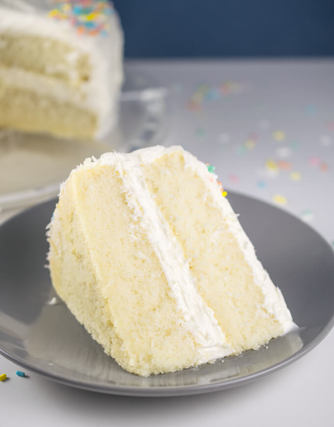 A slice of vegan white cake on a plate, showing tall, fluffy layers of pale golden cake between layers of white frosting.