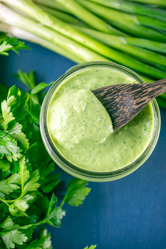 Overhead view of a jar of vegan green goddess dressing with a wooden spoon being dipped into it, surrounded by parsley and scallions