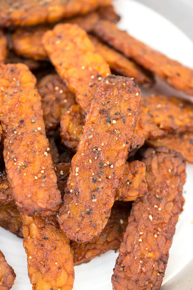 Closeup of a pile of many strips of tempeh bacon on a white plate.