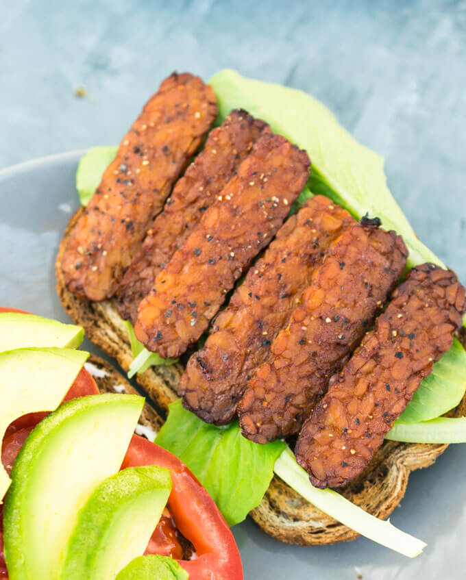 Six slices of tempeh bacon sprinkled with black pepper on top of a piece of bread with lettuce