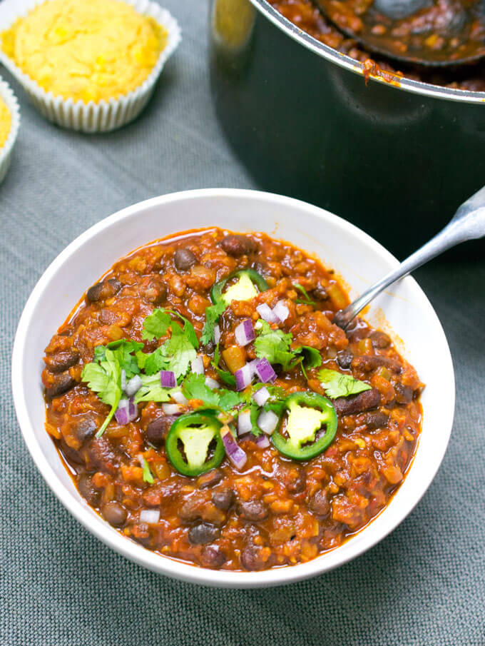 A bowl of vegan chili, showing a rich red color and thick texture. The bowl is garnished with fresh cilantro, red onions, and jalapeno slices.