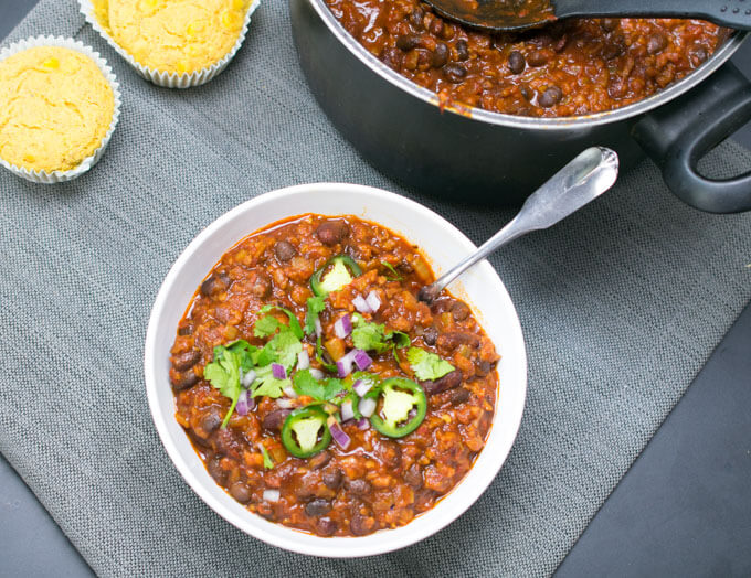 A bowl of vegan chili next to a full pot of the chili in the background along with two cornbread muffins