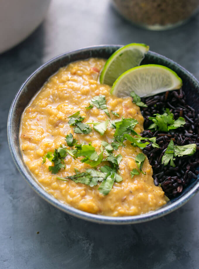 A bowl of creamy coconut lentil curry served in a blue ceramic bowl with wild rice. The curry is garnished with two lime wedges and a handful of fresh cilantro.