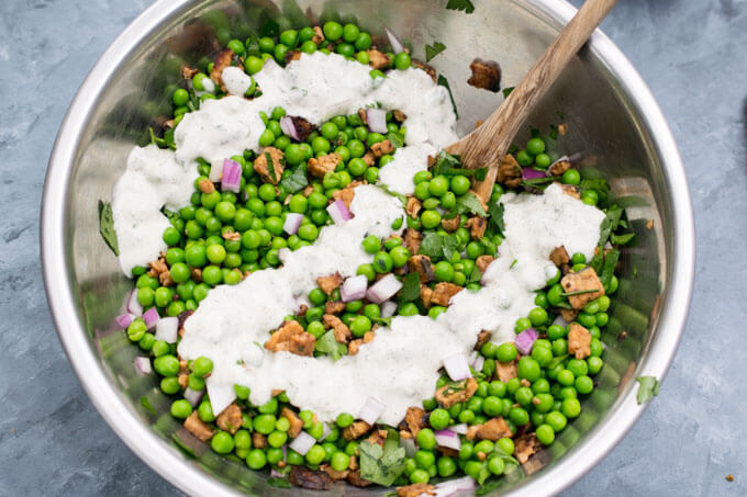 Vegan ranch dressing being mixed into a metal bowl filled with green peas, diced red onion, and browned tempeh bits