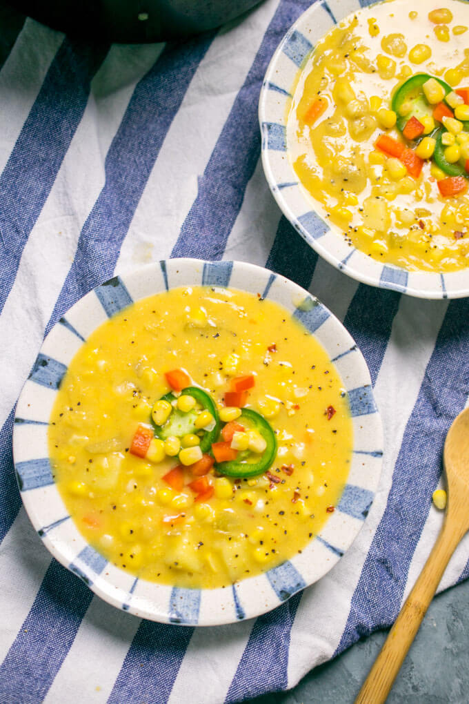 Two bowls of vegan corn chowder next to a wooden spoon, garnished with jalapeno and chopped bell pepper.