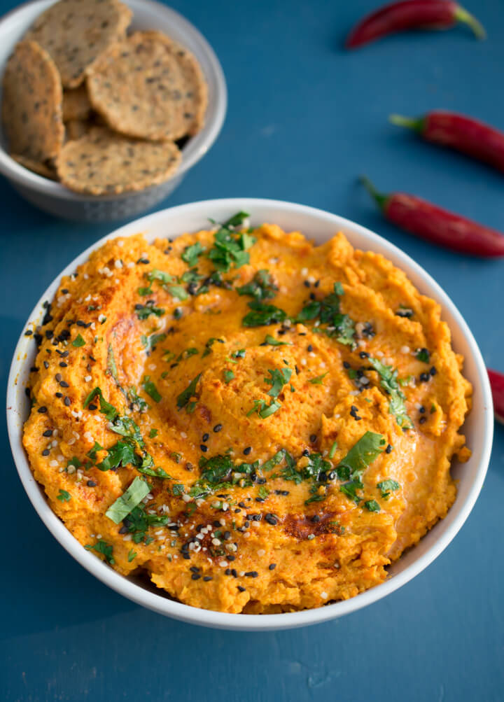 Roasted carrot hummus in a while bowl, garnished with cilantro with a small bowl of crackers in the background
