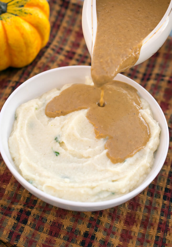 Brown gravy being poured onto a bowl of vegan cauliflower mash with a decorative gourd in the background.