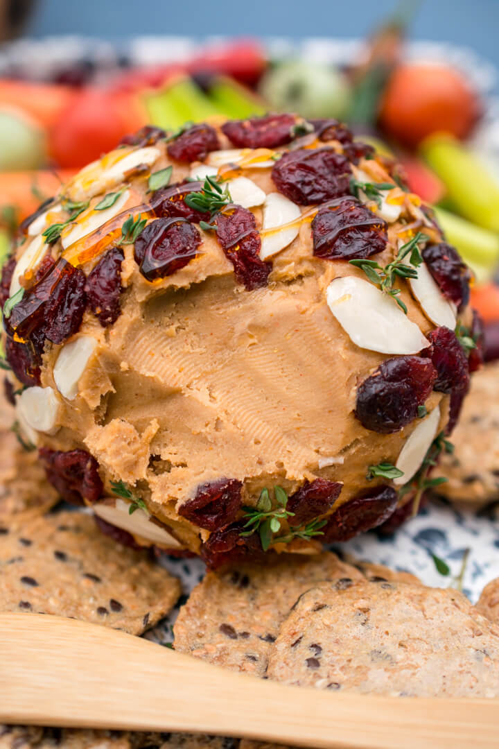 An orange hued vegan cheese ball decorated with dried cranberries and almonds and drizzled with honey