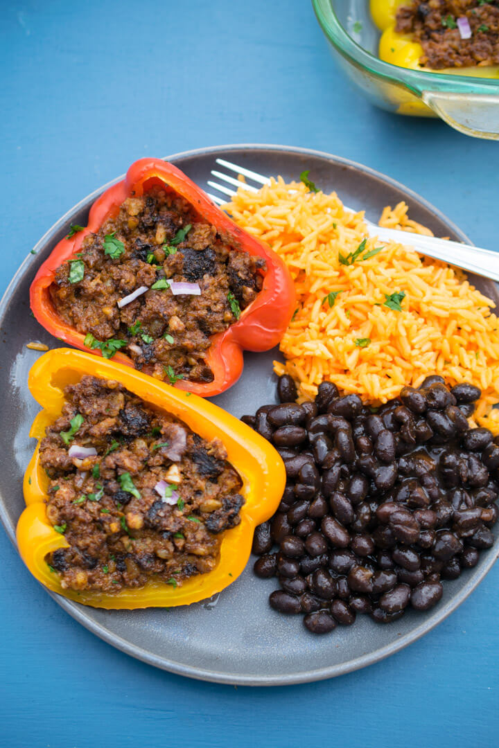 Two vegan stuffed peppers arranged on a plate next to some rice and beans.